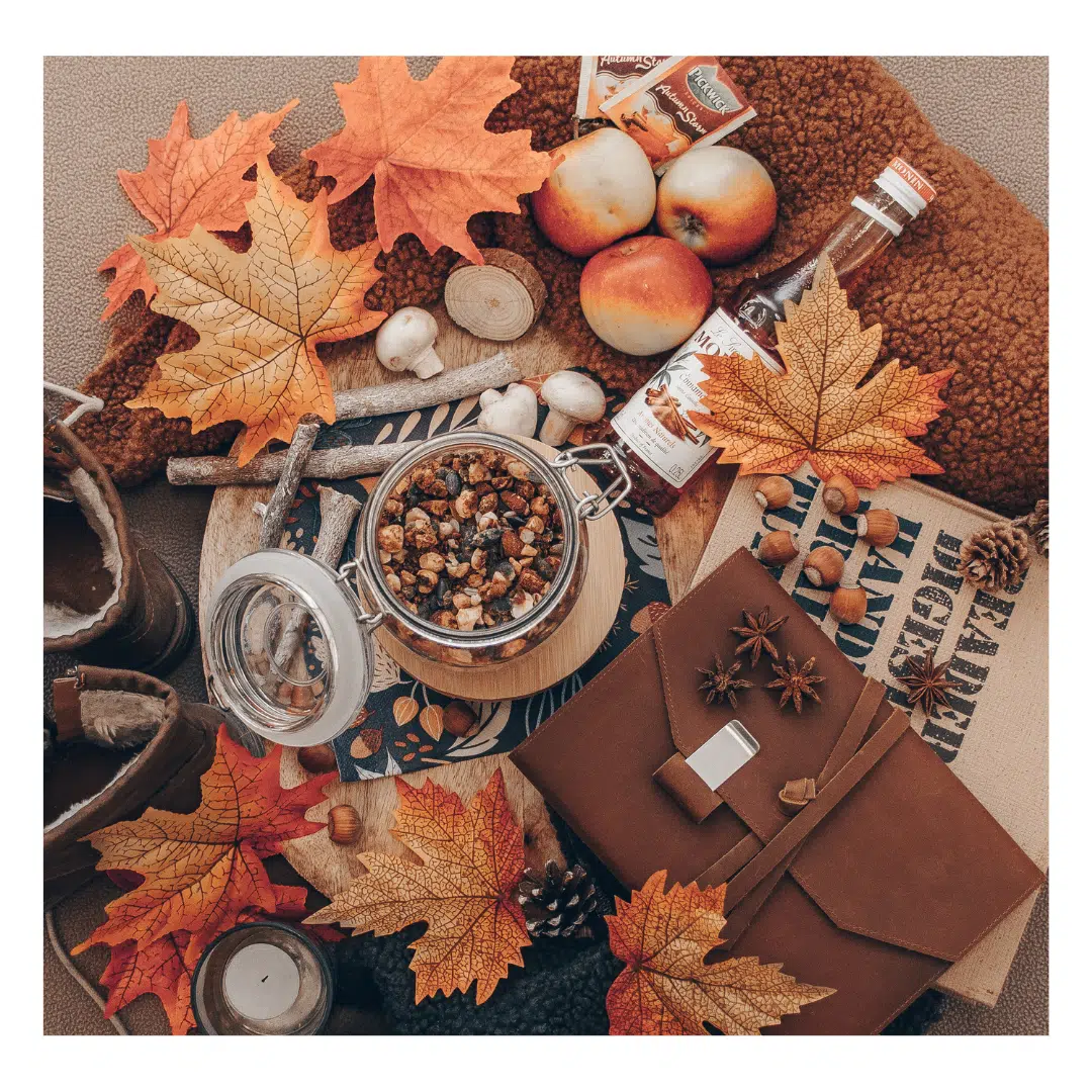 Een moodboard met herfstthema op een lichte achtergrond. Te zien zijn herfstbladeren, appels, een glazen pot met noten en gedroogd fruit, een fles, een leren notitieboekje, en enkele andere herfstgerelateerde items.