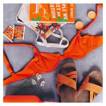 Een moodboard met zomerse items op een grijze achtergrond. Te zien zijn een oranje bikini, een paar oranje sandalen, een flesje water, een boek met een oranje omslag, twee theezakjes, een canvas tas en enkele bloemen.