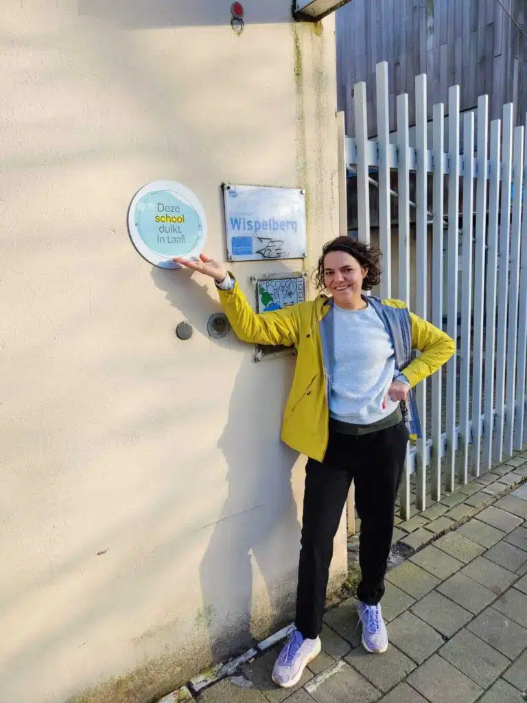 Vrouw in geel jasje wijst trots naar het 'Deze school duikt in taal!'-logo en het naambord van basisschool Wispelberg