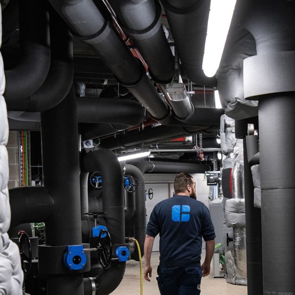 Imtech-technicus loopt door machinekamer met zwart geïsoleerde leidingen en blauwe kleppen tijdens fotoshoot voor Graviteit