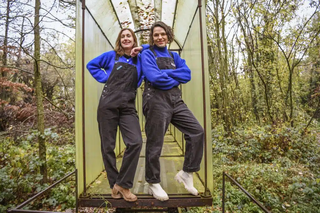 Twee vrouwen van Graviteit in matching blauwe sweaters en zwarte tuinbroek poseren in een glazen serre omringd door herfstbom