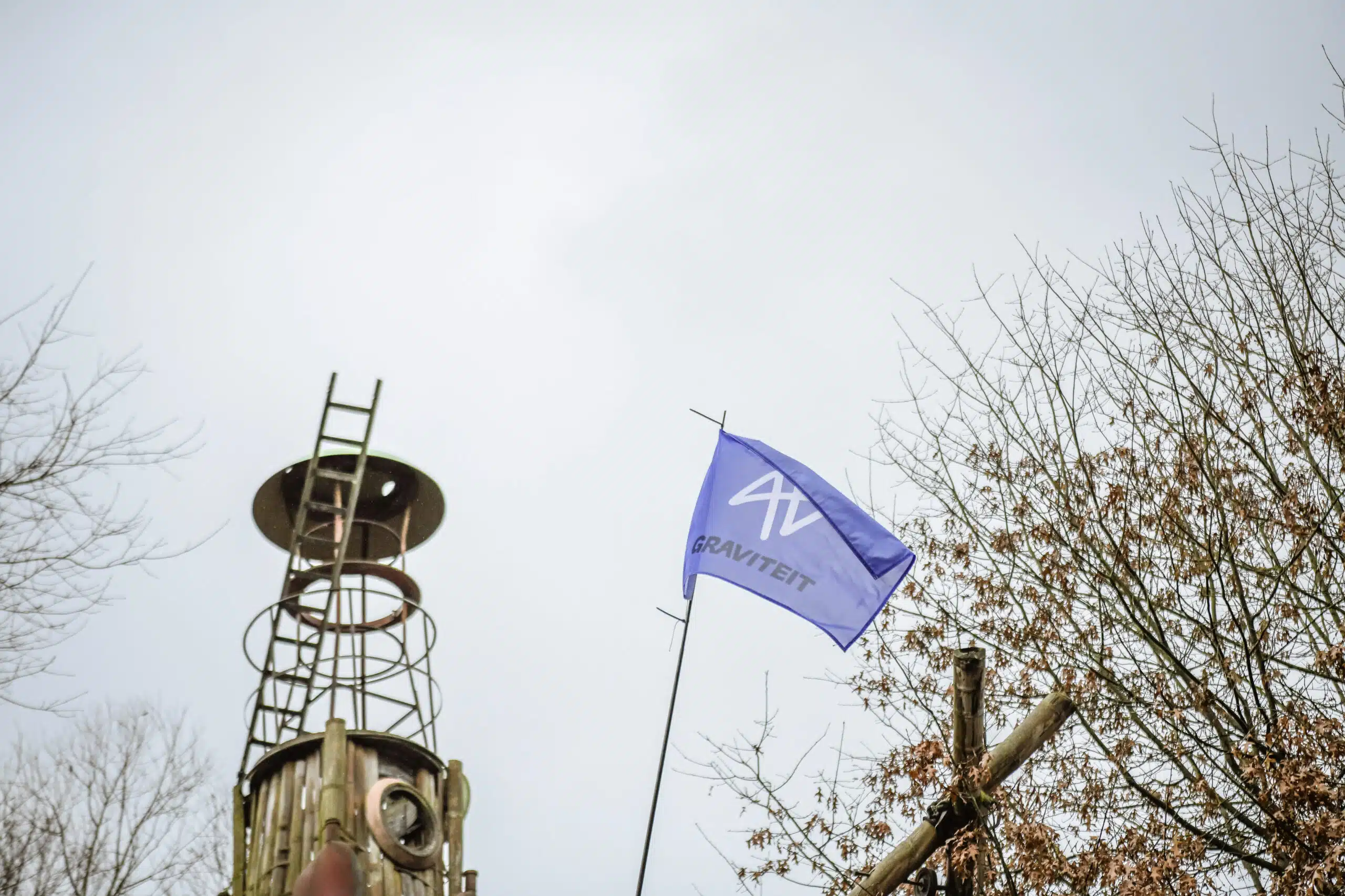 Graviteit-vlag wapperend aan een mast naast een industriële metalen toren en kale bomen, tijdens de rebranding fotoshoot