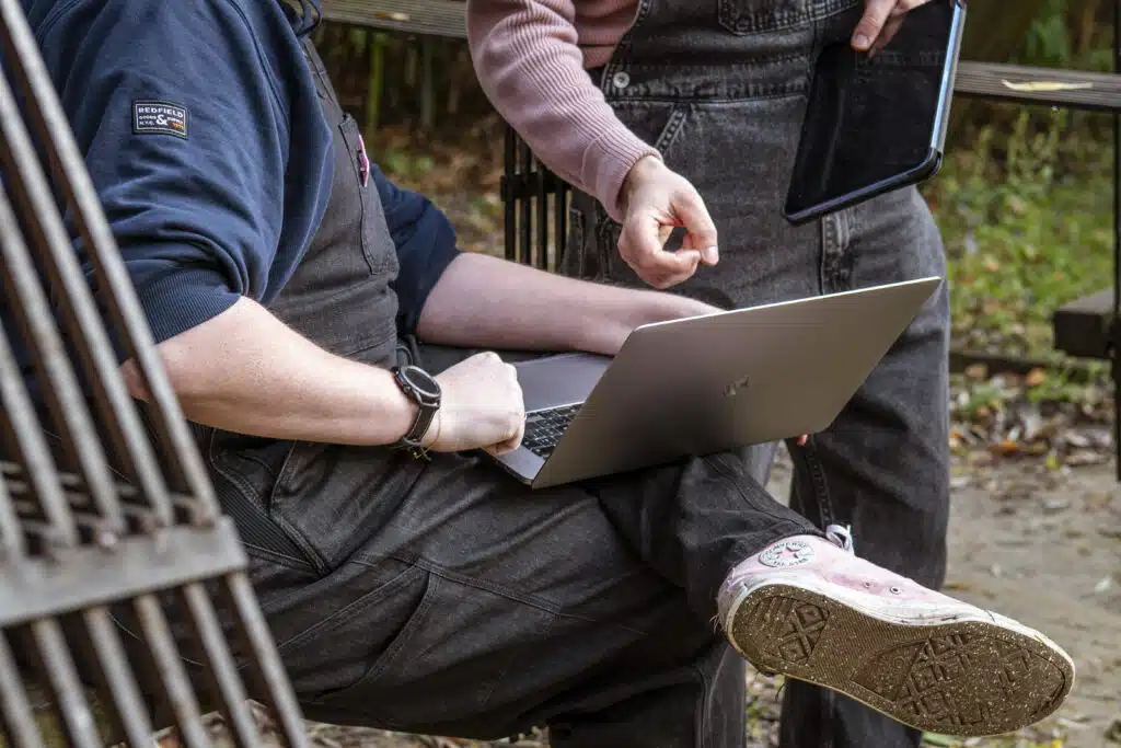 Twee personen buiten op een bankje die samenwerken aan copywriting, met een laptop en tablet in handen, omgeven door groen.