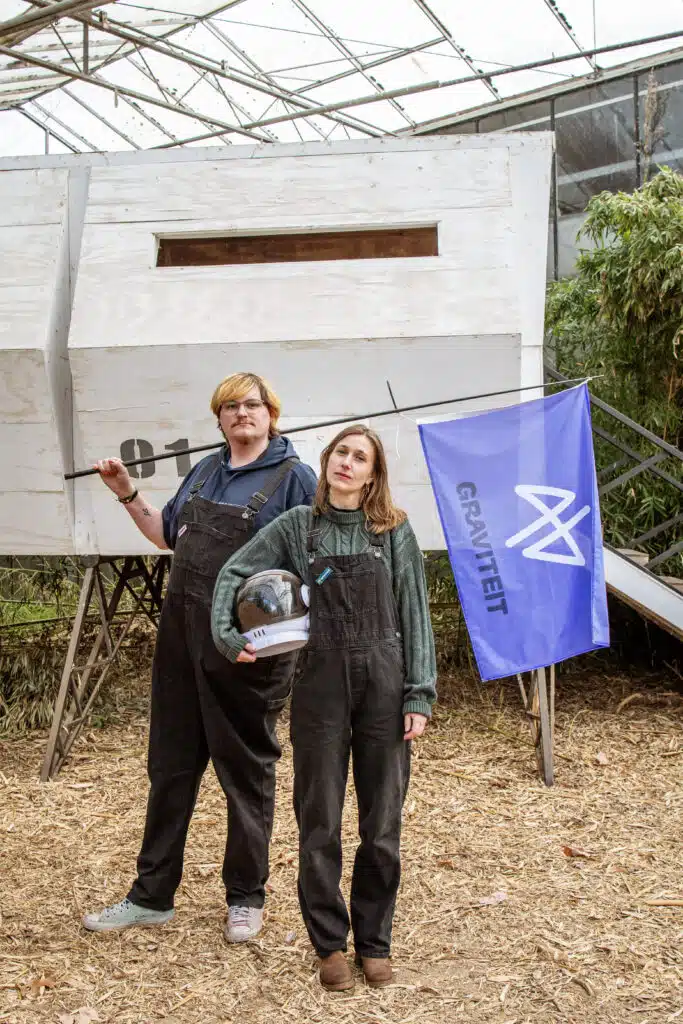 Twee Graviteit-teamleden in tuinbroek poseren voor een houten installatie in een serre, met een paarse Graviteit-vlag
