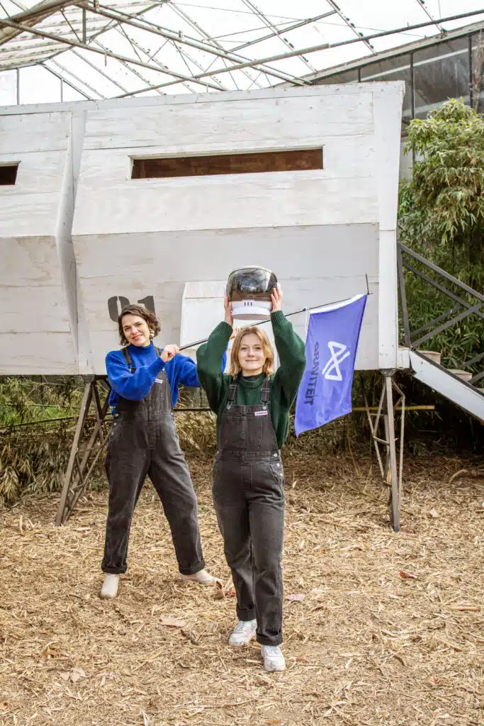 Twee Graviteit-teamleden in tuinbroek poseren enthousiast in een industriële ruimte met Graviteit-vlag op de achtergrond.