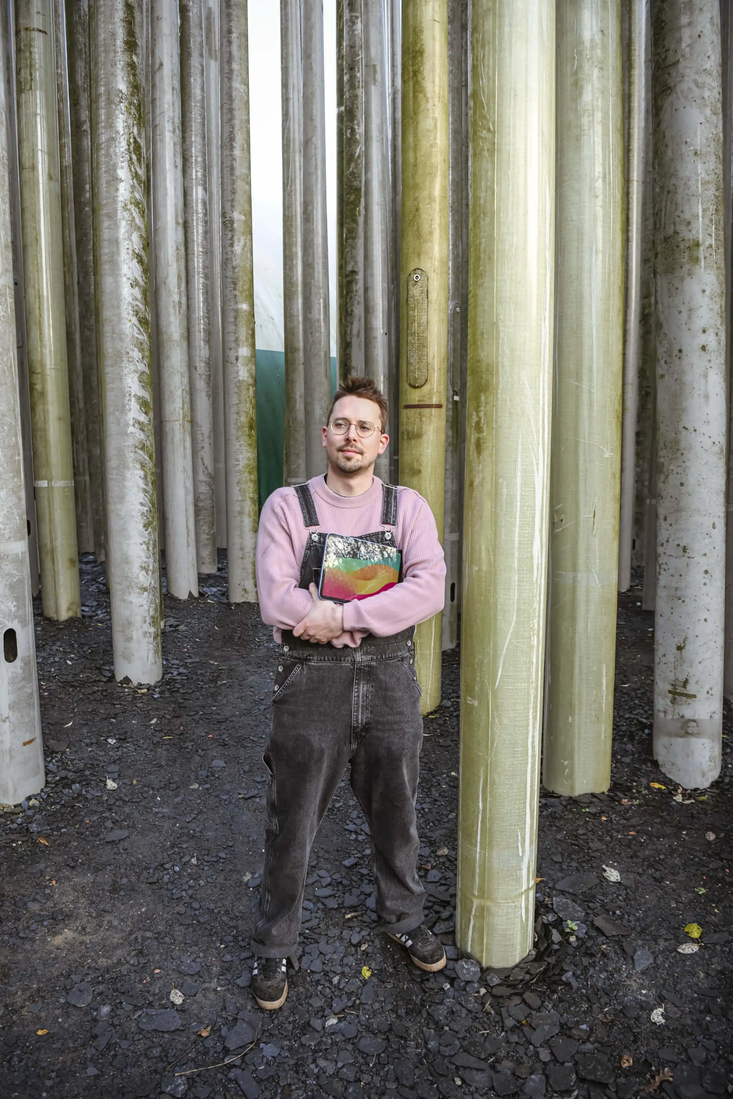 Graviteit-teamlid in roze trui en grijze overall met een boek in de hand, staand tussen industriële zuilen voor een creatief