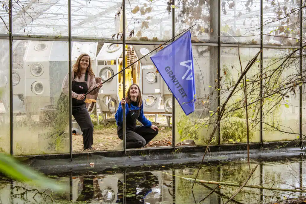 Twee Graviteit-medewerkers met een blauwe Graviteit-vlag in een verlaten serre, omgeven door industriële objecten en waterref