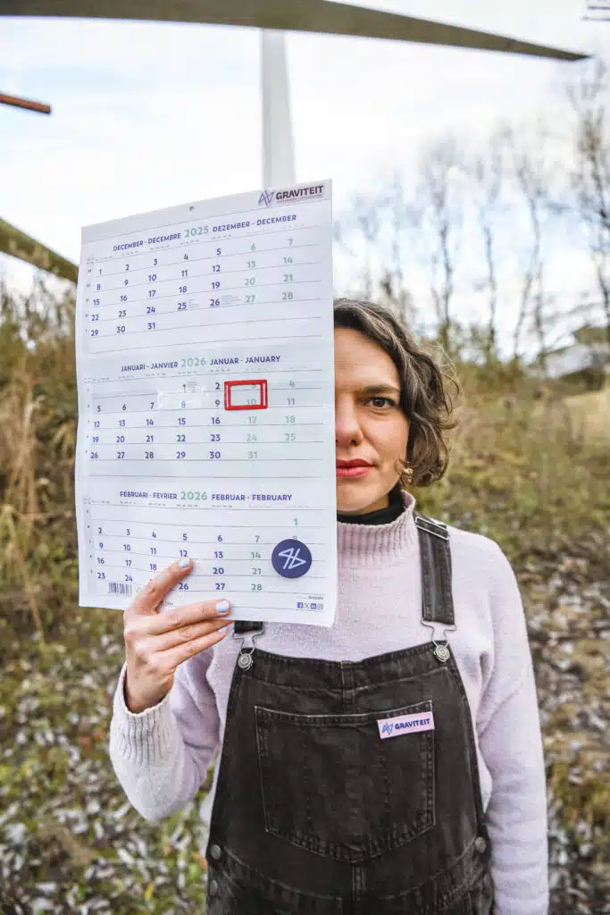 Graviteit teamlid in tuinbroek houdt een Graviteit-kalender 2025-2026 vast met januari 2026 omcirkeld, buiten gefotografeerd