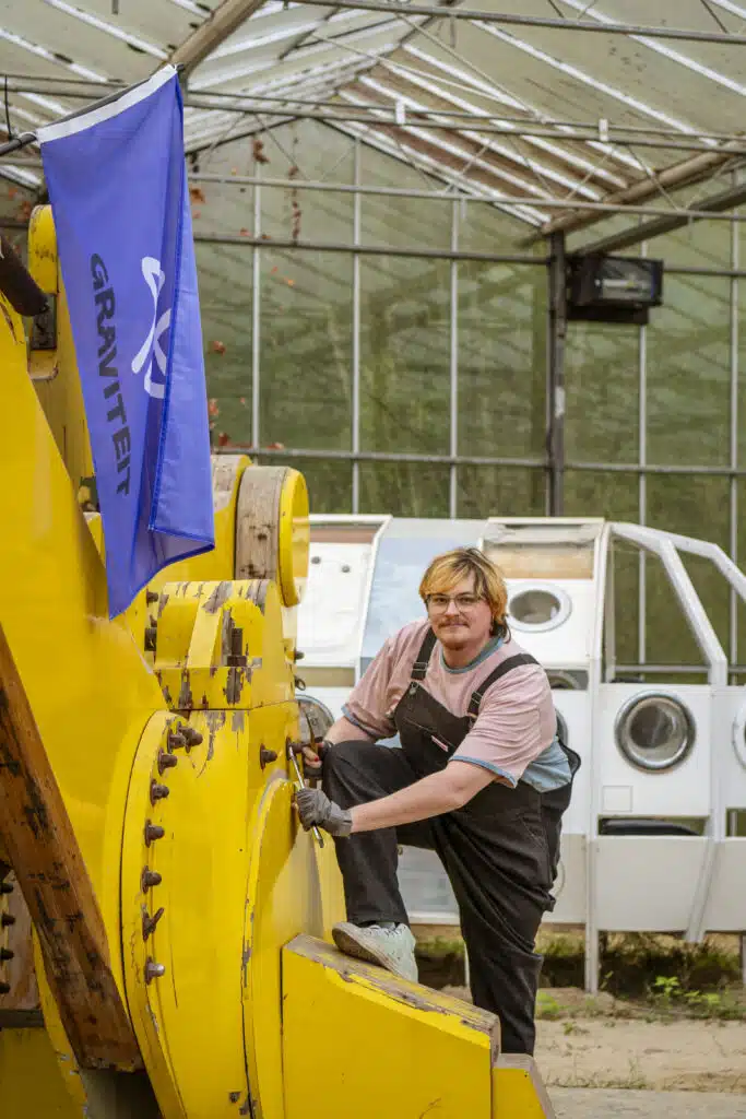 Graviteit teamlid in tuinoverall sleutelt aan gele graafmachine in serre, met Graviteit-vlag – creatief communicatiebureau