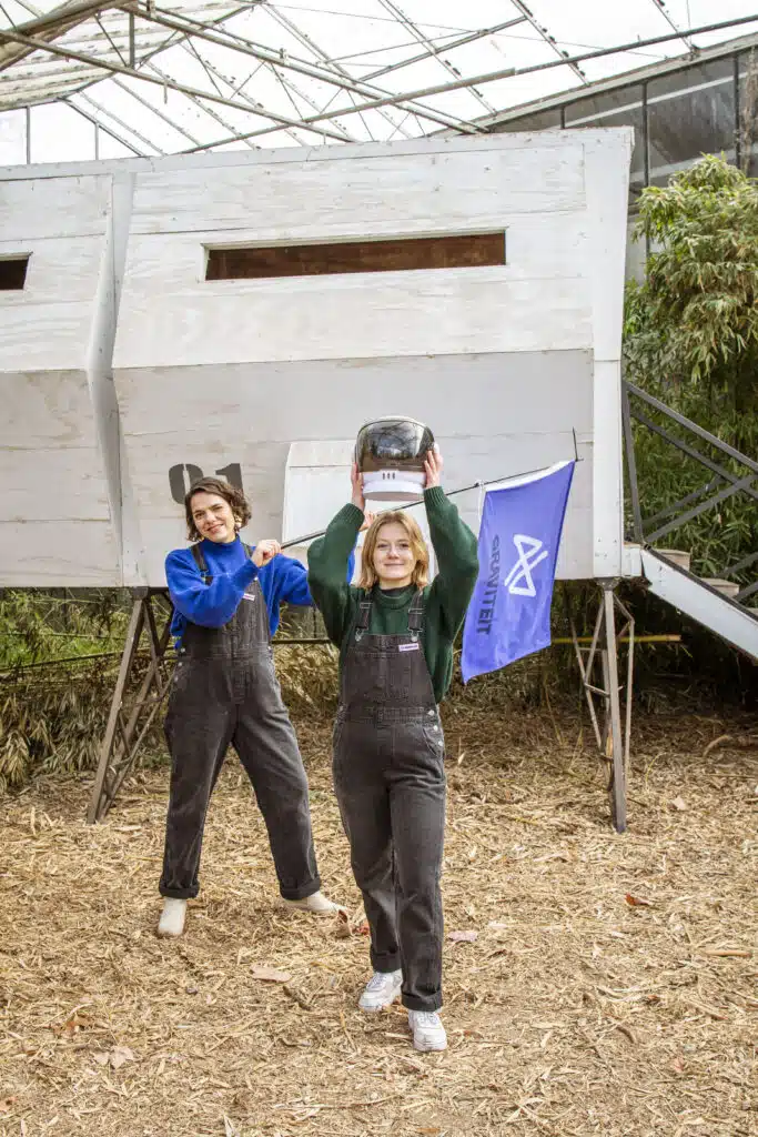 Twee teamleden van Graviteit in tuinbroek poseren voor een houten constructie in een serre, met het Graviteit-logo op een vla
