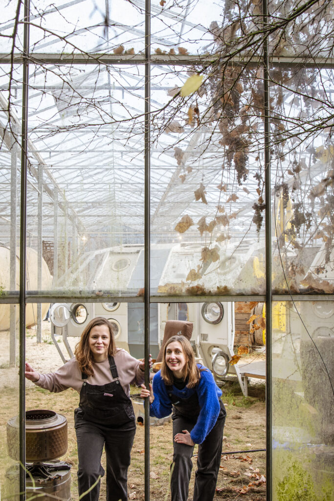 Twee vrouwen van het creatieve team van Graviteit poseren glimlachend in een glazen serre vol vintage objecten tijdens een re