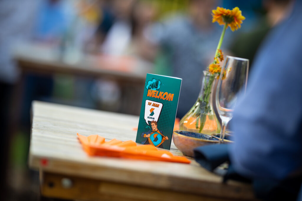 Een uitnodigingskaartje voor een evenement van Sirus om hun 10-jarig bestaan te vieren met de tekst "Welkom" op de voorgrond, op een houten tafel naast een bloemenvaas en glazen.