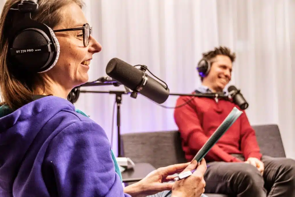 Twee mensen nemen een podcast op in een studio, vrouw met paarse hoodie spreekt in microfoon, man luistert op achtergrond