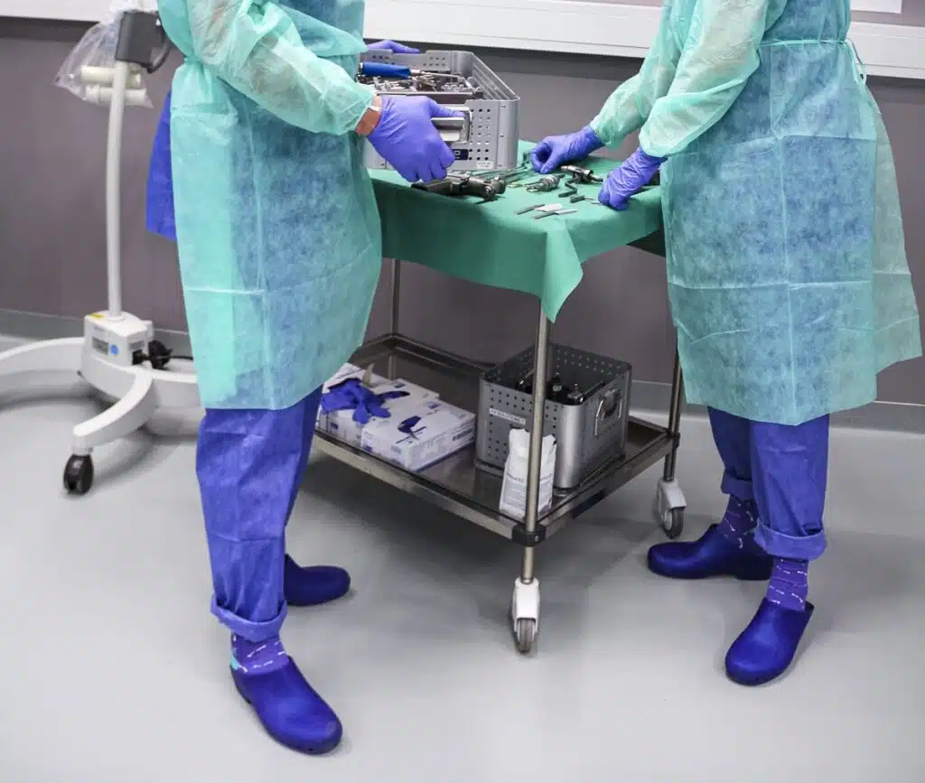 Twee medische professionals bereiden chirurgische instrumenten voor op een mobiele werkplek in een klinische setting. Ze zijn gekleed in beschermende schorten, handschoenen, klompen en BeMedico sokken, wat zorgt voor een steriele omgeving. De focus ligt op hun nauwkeurige voorbereiding, wat het belang van hygiëne en precisie in medische procedures benadrukt.