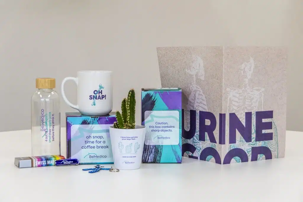 Een verzameling promotieartikelen van BeMedico, waaronder een glazen fles met houten dop, een witte mok met de tekst "OH SNAP!", een doos met de tekst "Caution, this box contains sharp objects.", een cactus in een potje met de tekst "I love how prickly your legs feel... said no one ever", een blauwe sleutelhanger in de vorm van een medisch implantaat, en een achtergrond met de tekst "URINE GOOD".
