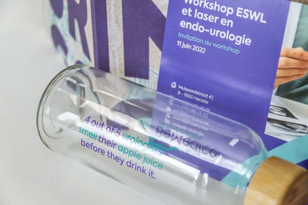 Een close-up met gadgets en flyers van BeMedico op de voorgrond is een glazen fles met een houten dop te zien, liggend voor een promotieflyer. De tekst op de fles luidt: "4 out of 5 urologists smell their apple juice before they drink it." Op de achtergrond is een paarse flyer te zien met informatie over een workshop endo-urologie.