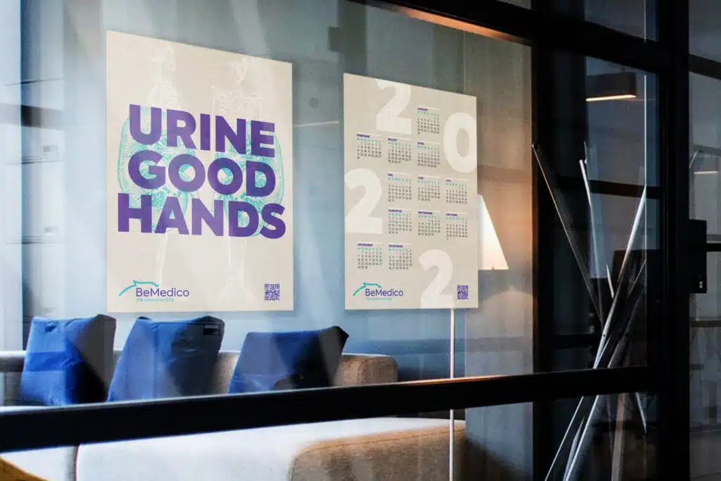Twee posters van BeMedico aan een glazen wand, gezien van buiten een kamer. De linkerposter heeft de tekst "URINE GOOD HANDS" en de rechterposter toont een kalender.