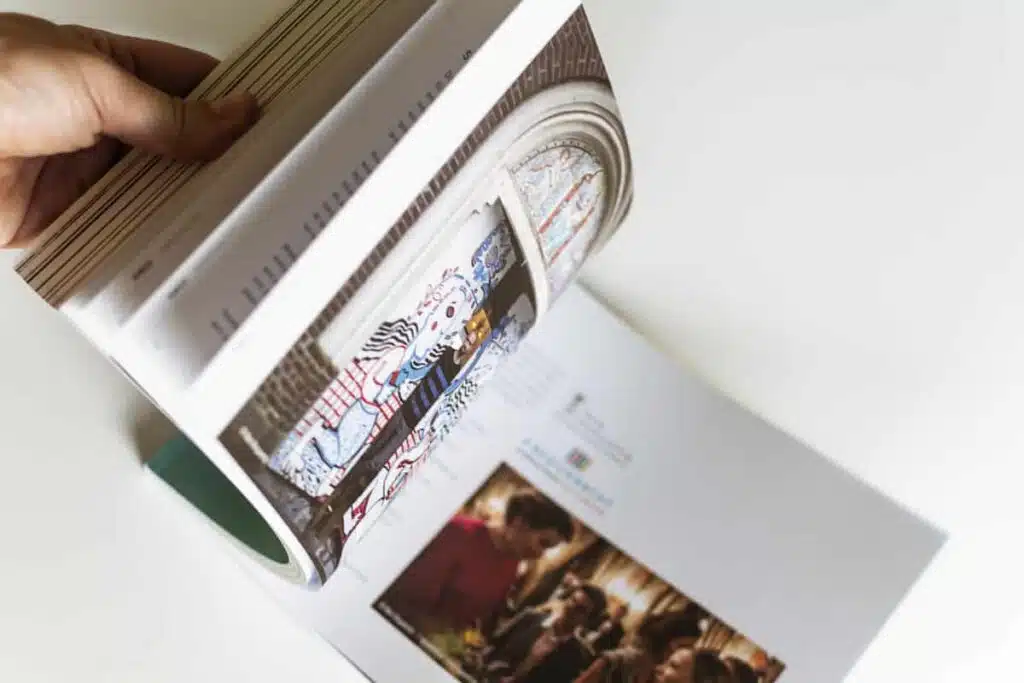 Een close-up foto van iemand die door het gedrukte jaarverslag van Veneco bladert, waarbij een foto van een gebrandschilderd raam en een foto van een groep mensen zichtbaar zijn.