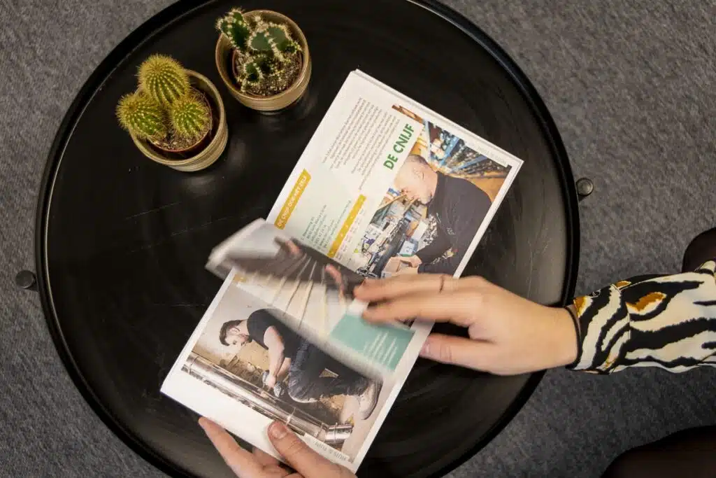 Magazine voor Winkelierde op tafel met cactussen: Een persoon bladert door een magazine met een zwarte tafel en cactussen op de achtergrond.
