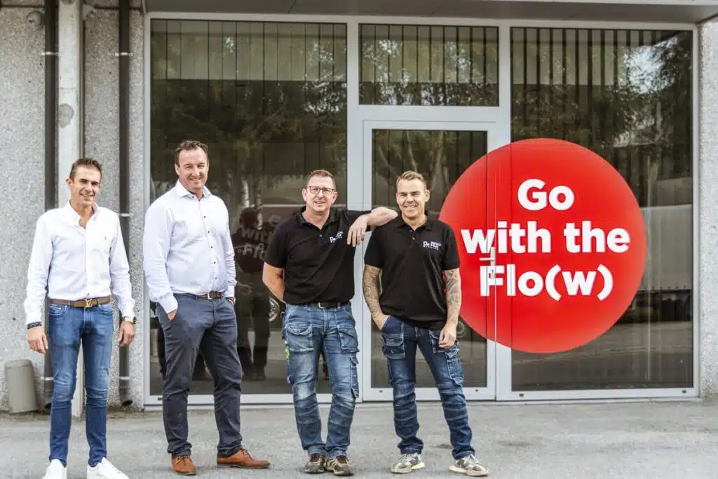 Vier medewerkers van Chapewerken De Flo poseren voor hun kantoor naast het rode logo met de slogan 'Go with the Flo(w)'