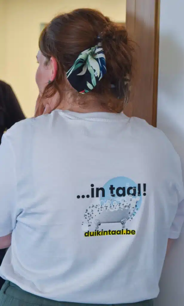 Een foto met de achterkant van een persoon die een wit T-shirt draagt met de tekst "...in taal!" en de URL "duikintaal.be".