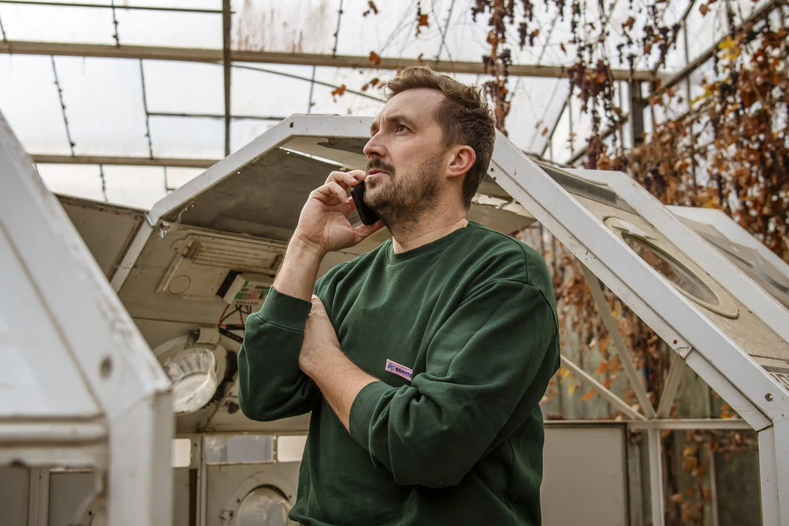 Graviteit-teamlid in groen sweatshirt belt met smartphone in een serre, bezig met communicatieplanning voor een klant.