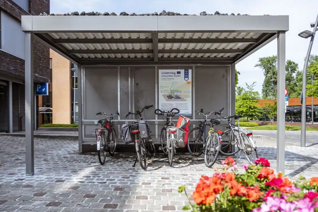 Een fietsenstalling met meerdere fietsen erin geparkeerd, met een informatiebord over het Interreg Cool Towns-project.