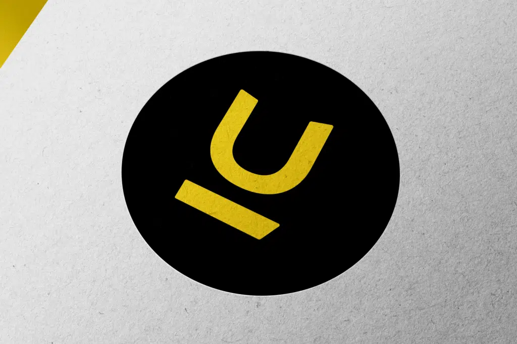 Een abstract logo van een gestileerde letter 'U' in geel op een zwarte cirkel, geplaatst tegen een lichtgrijze achtergrond.