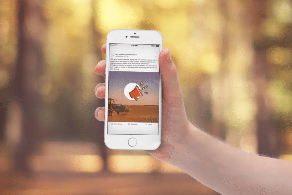 Hand die een smartphone vasthoudt waarop een social media post van het Vlaams Agrarisch Centrum (VAC) zichtbaar is, met een achtergrond van landbouwgrond.