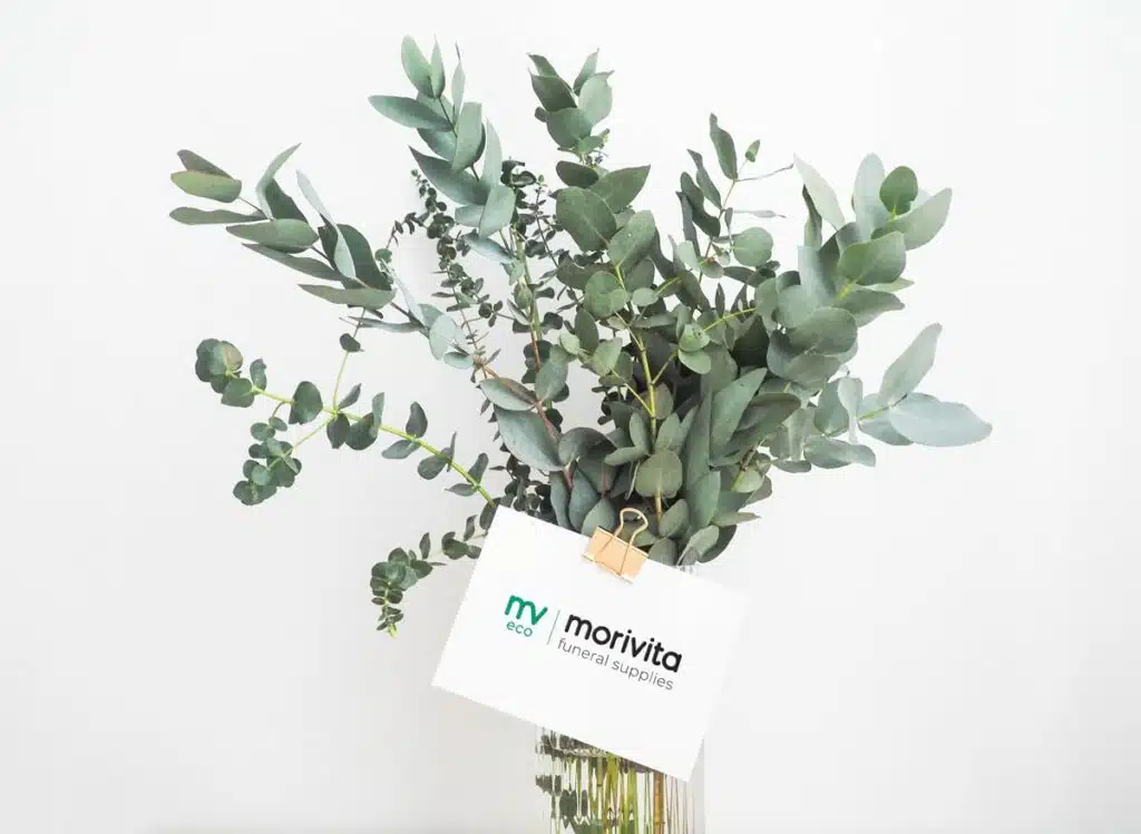 Eucalyptusboeket met Morivita-eco kaart: Een boeket met groene eucalyptusbladeren in een glazen vaas, waaraan een kaartje met het logo van Morivita funeral supplies is vastgemaakt met een goudkleurige clip.