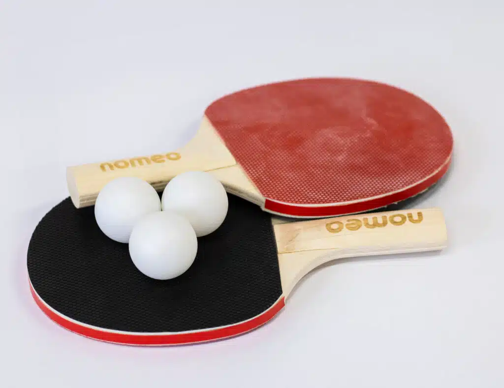 Twee tafeltennisbatjes, één met een rode en één met een zwarte kant, met drie witte pingpongballen en het logo "nomeo" op de handvatten.