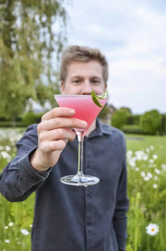 Een man in de buitenlucht die een roze cocktail in een martiniglas naar de camera houdt.