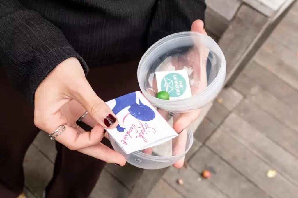 Vrouwenhanden houden een transparante ronde doos vast met daarin een kaartje, sticker en groen snoepje — Sjiek Verdriet campa