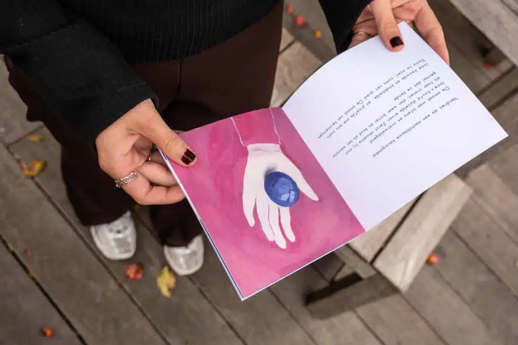 Persoon houdt een open boekje vast met illustratie van een hand die een blauwe steen vasthoudt — grafisch ontwerp voor Sjieke