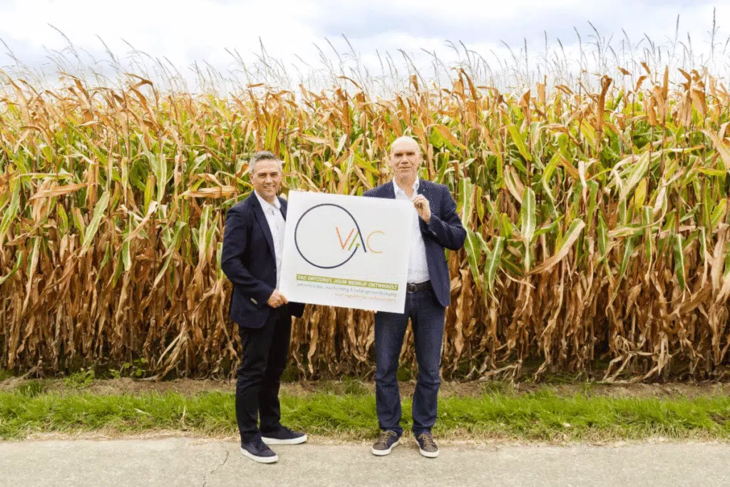 Twee mannen in pak poseren voor een veld met maïs, terwijl ze een bord vasthouden met het logo van het Vlaams Agrarisch Centrum (VAC).