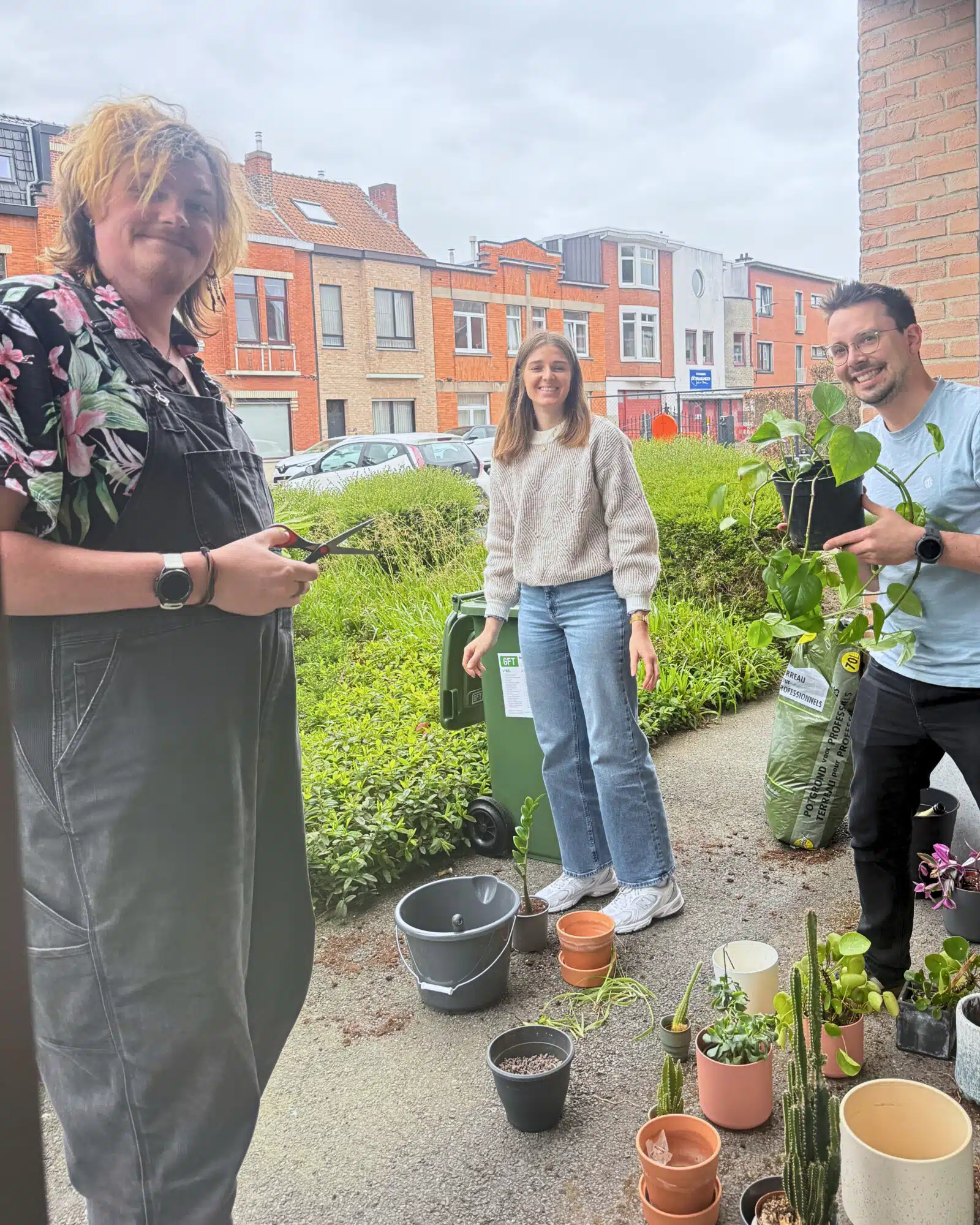 Drie mensen bezig met tuinieren en planten verzorgen in een stedelijke omgeving met kleurrijke huizen op de achtergrond