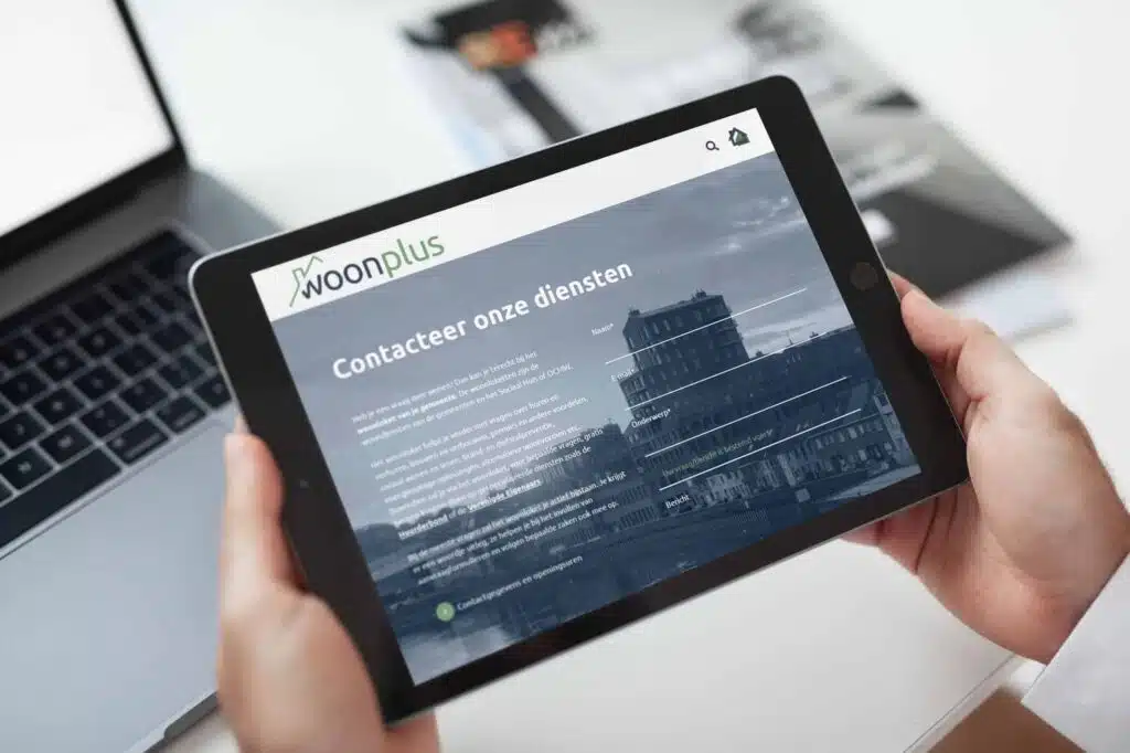 Close-up van een tablet die de contactpagina van de Woonplus website toont, waarbij een hand het apparaat vasthoudt en een laptop op de achtergrond staat. De pagina bevat een formulier voor contactinformatie.