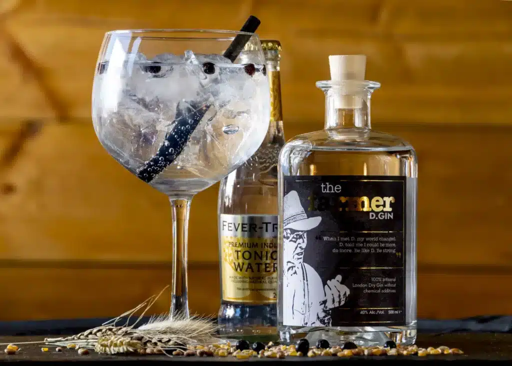 The Farmer D.GIN fles naast een gin-tonic cocktail in ballon glas met Fever-Tree tonic en zwarte rietje, omringd door graanko