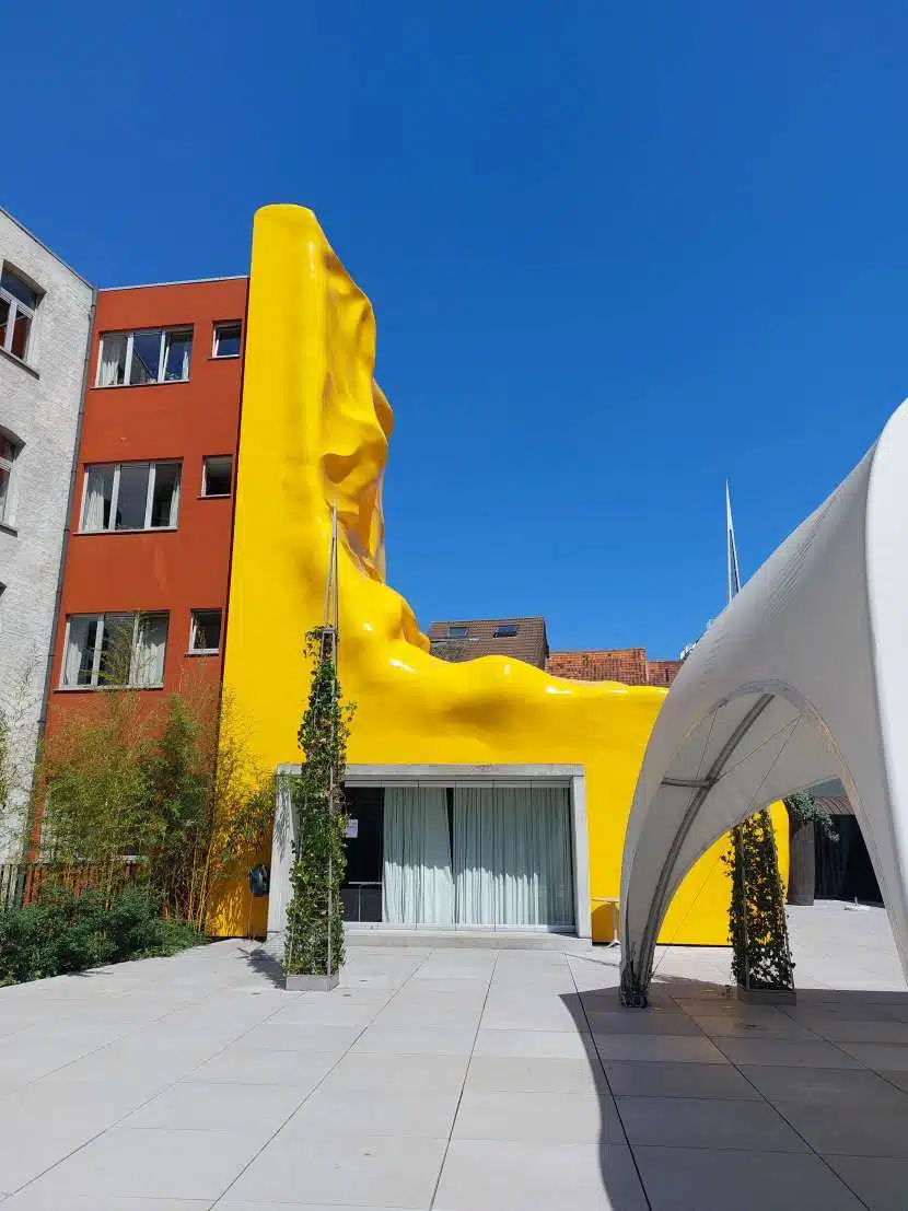 Gevel van een gebouw met een felgele, golvende structuur die tegen de zijkant van het gebouw is geplaatst, onder een heldere blauwe lucht.