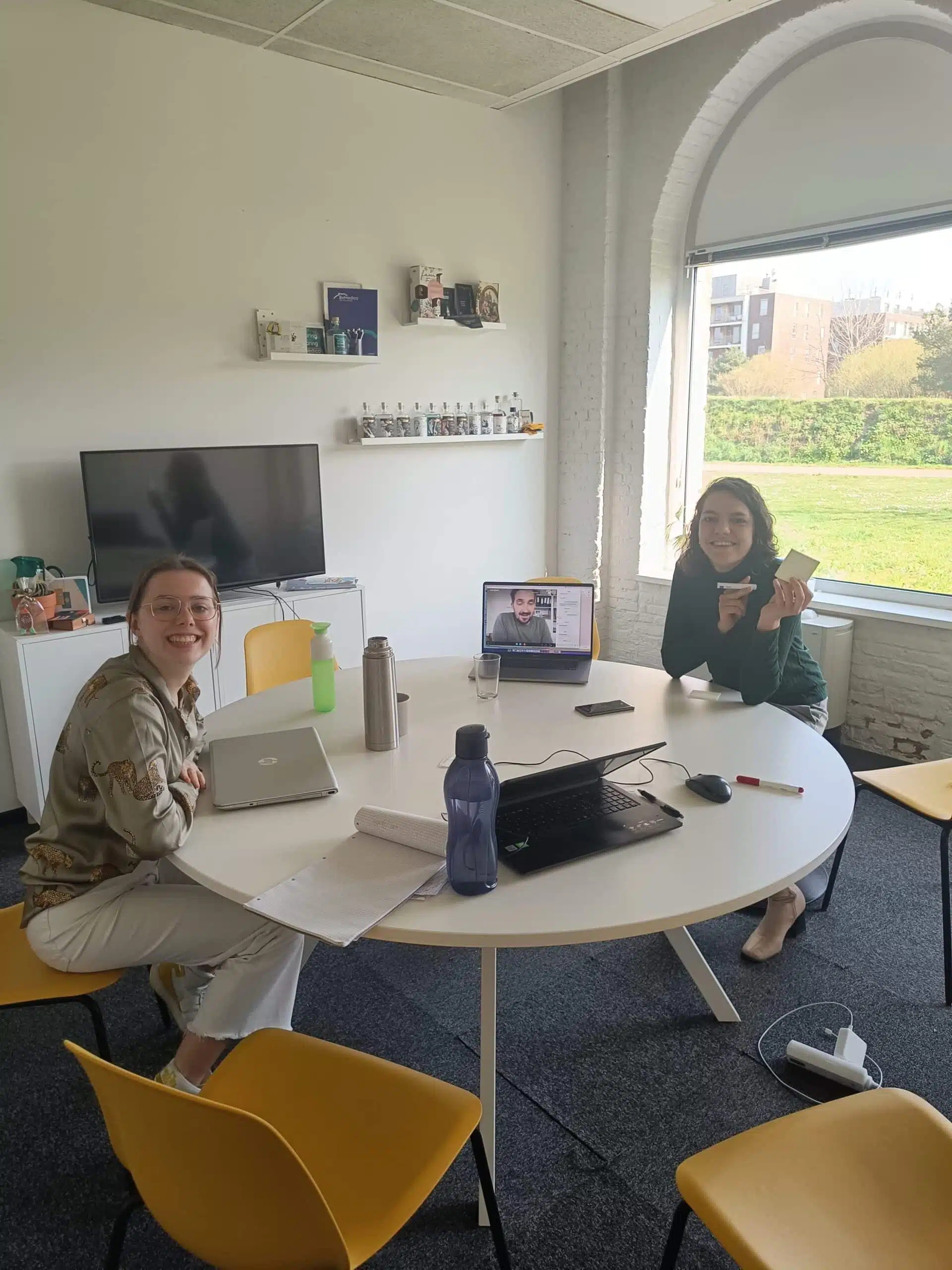 stagiaire bedrijfscommunicatie Bedrijfscommunicatie stagiair Julie en Ines van Graviteit zitten aan een ronde tafel in een vergaderruimte, Frank van Graviteit is zichtbaar via een scherm tijdens een online vergadering.