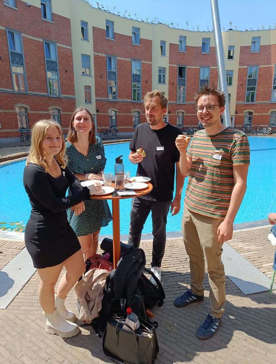 Het Graviteit team staat buiten rond een hoge tafel bij een zwembad, genietend van een maaltijd in de zon. De achtergrond toont een rood bakstenen gebouw met grote ramen.