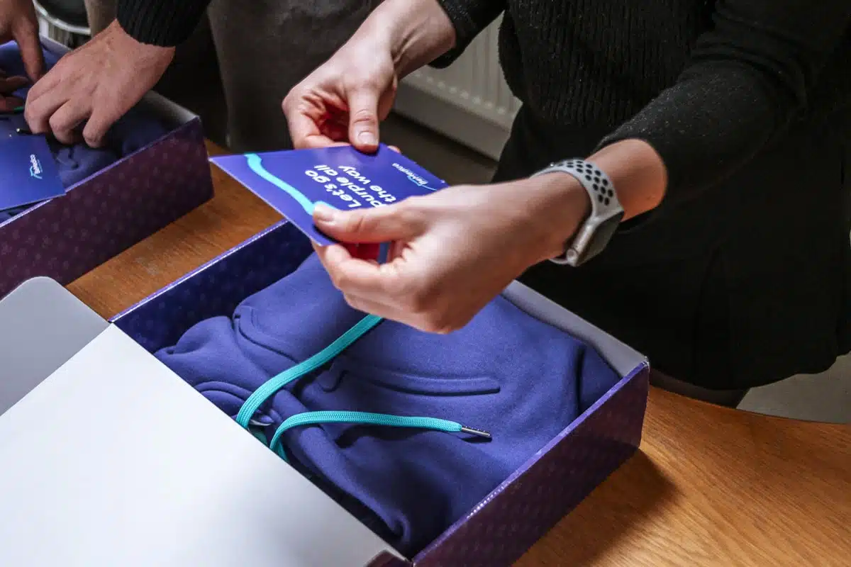 Iemand is een paarse hoodie met blauwe trekkoorden aan het uitpakken. Er is een kaart zichtbaar met de slogan "Let's go purple all the way." Dit was voor de eindejaarscampagne van BeMedico.