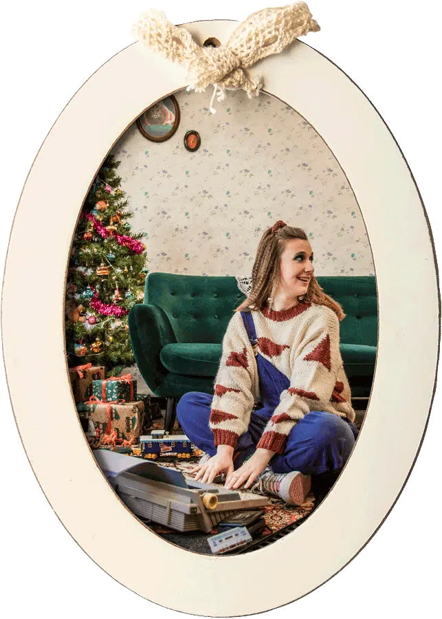 Febe van Graviteit zittend op de grond in een retro outfit, spelend met een vintage spelcomputer voor een kerstboom, ingelijst in een ovale lijst met een strik bovenaan.