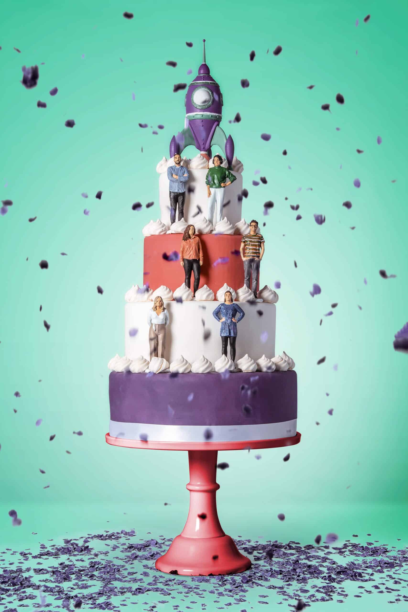 Een grote, feestelijke taart met drie lagen in paars, rood en wit. Bovenop de taart staat een raket en de verschillende lagen zijn versierd met miniatuursculpturen van het Graviteit team die elk op een andere laag van de taart staan. Paarse confetti valt om de taart heen, wat de feestelijke sfeer versterkt.