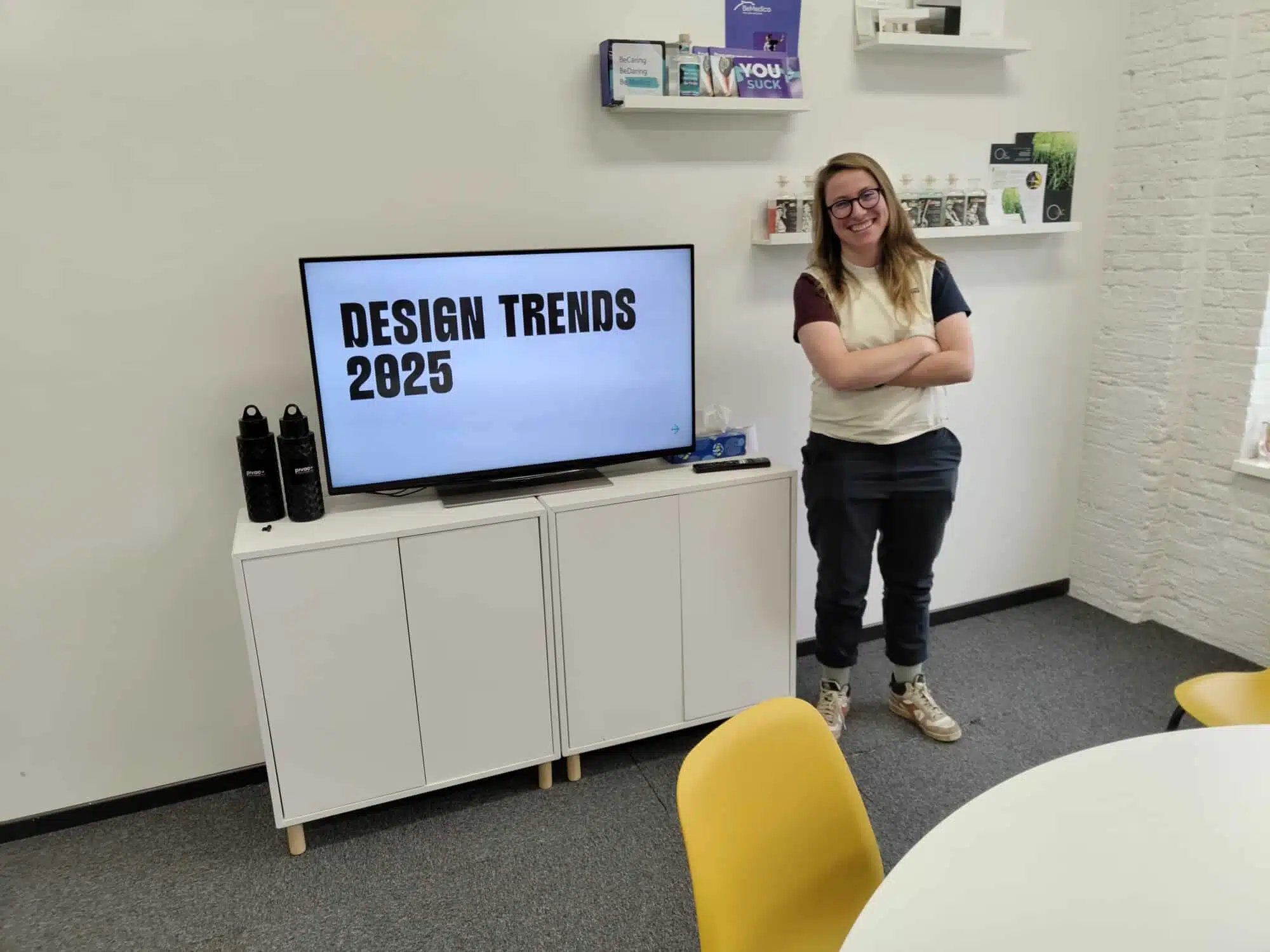 Stagiaire Jana poseert naast tv-scherm met "Design Trends 2025" in modern kantoor met witte muren en gele stoelen