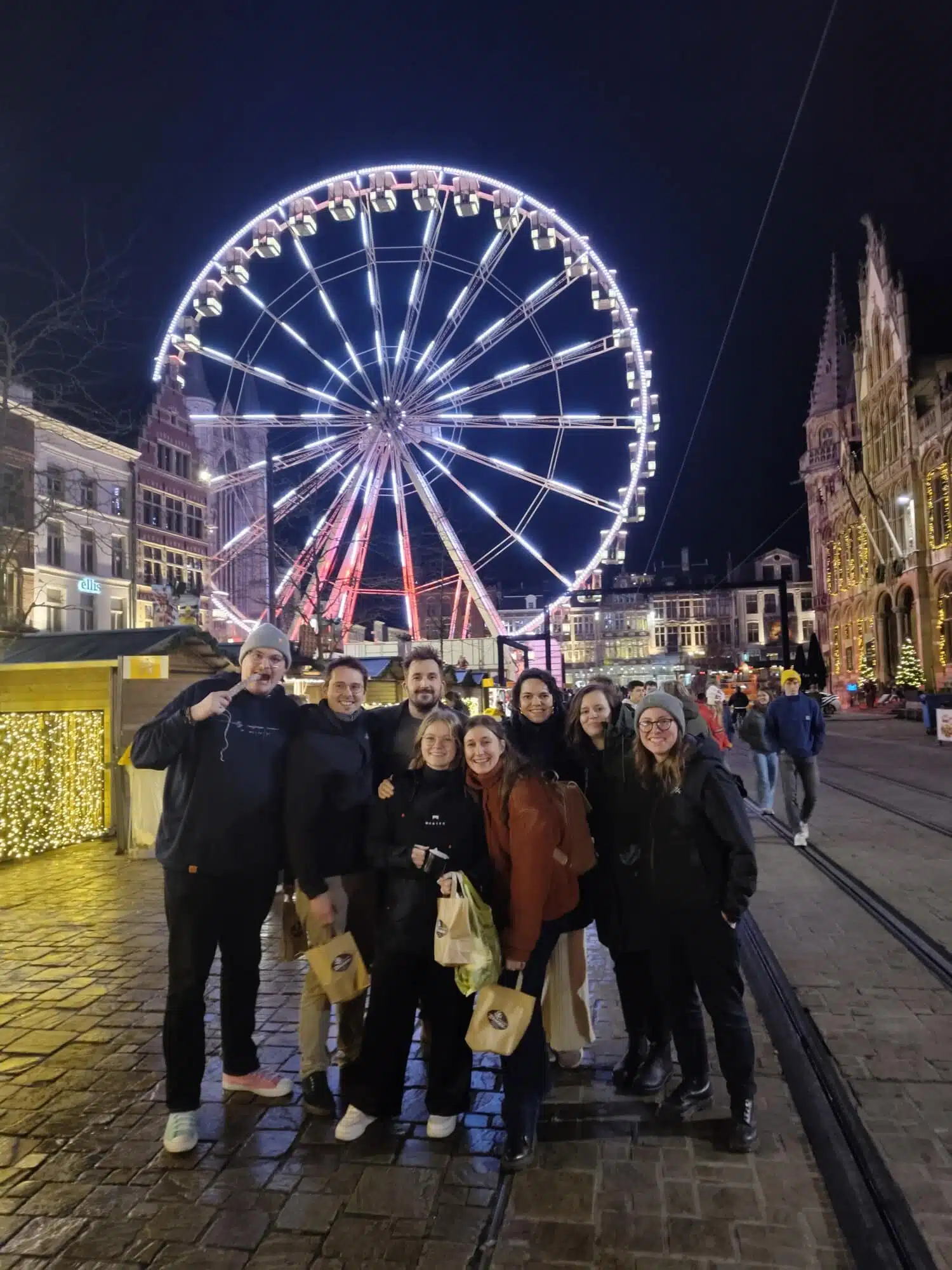 Groep vrienden poseert 's avonds voor verlicht reuzenrad op historisch stadsplein tijdens winteruitje