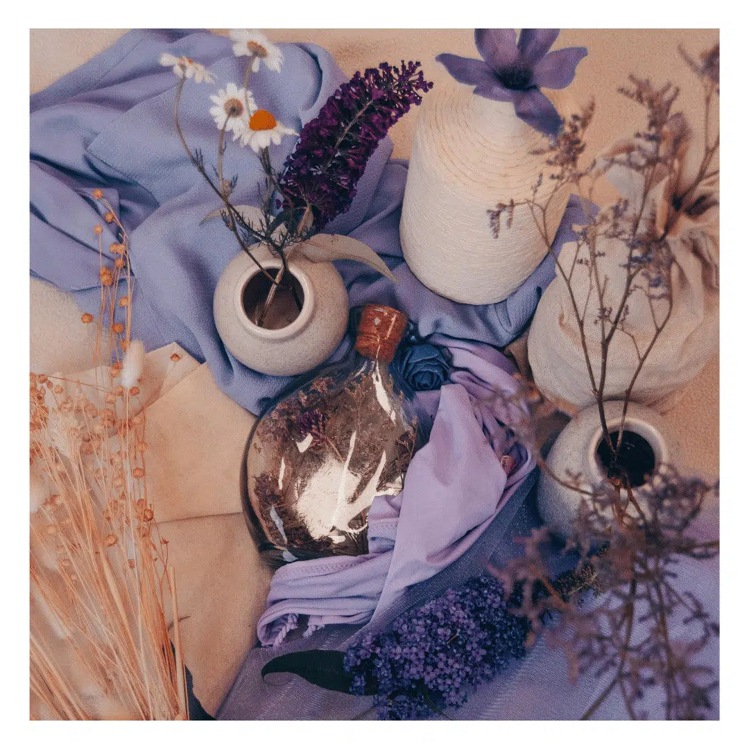 Een moodboard met paarse en beige tinten op een lichte achtergrond. Te zien zijn gedroogde bloemen, een paarse sjaal, een decoratieve fles, vazen, en verschillende stoffen en decoraties.
