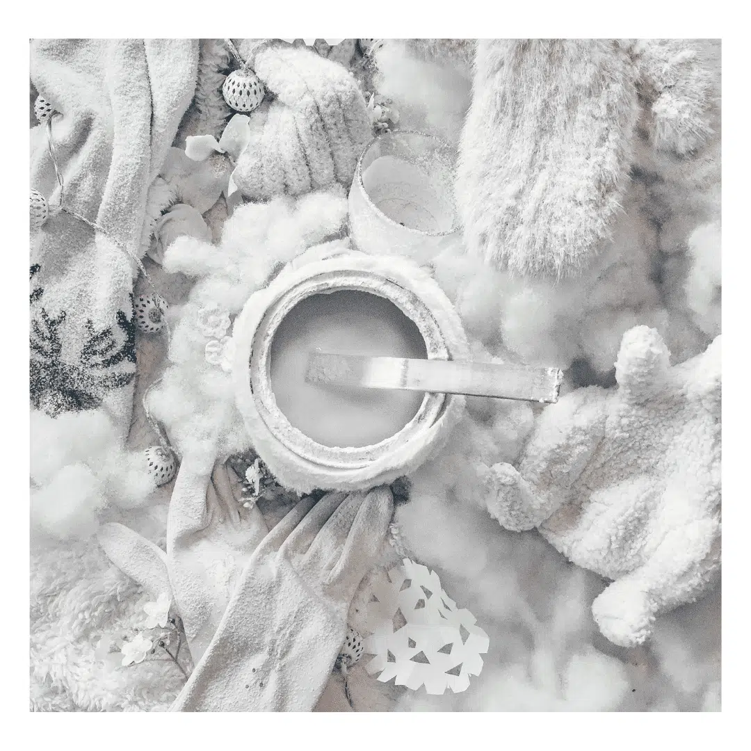 Een moodboard met een winterthema, voornamelijk in witte en grijze tinten. Te zien zijn een pot met verf en een kwast, warme kledingstukken zoals een wollen muts, handschoenen en een trui, en decoratieve elementen zoals sneeuwvlokken en kleine witte bolletjes.