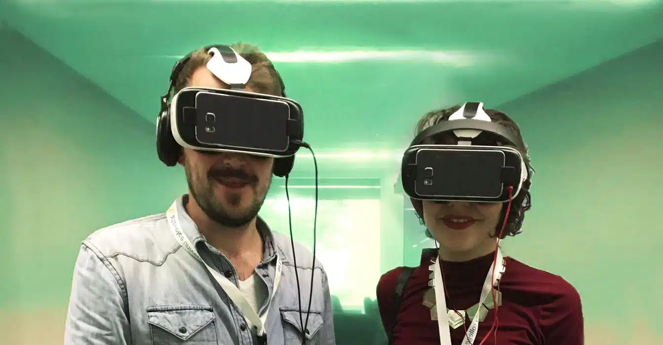 Frank Mertens en Ines Maroye met virtual reality-headsets en koptelefoons, lachend terwijl ze in een futuristische, groene ruimte staan.