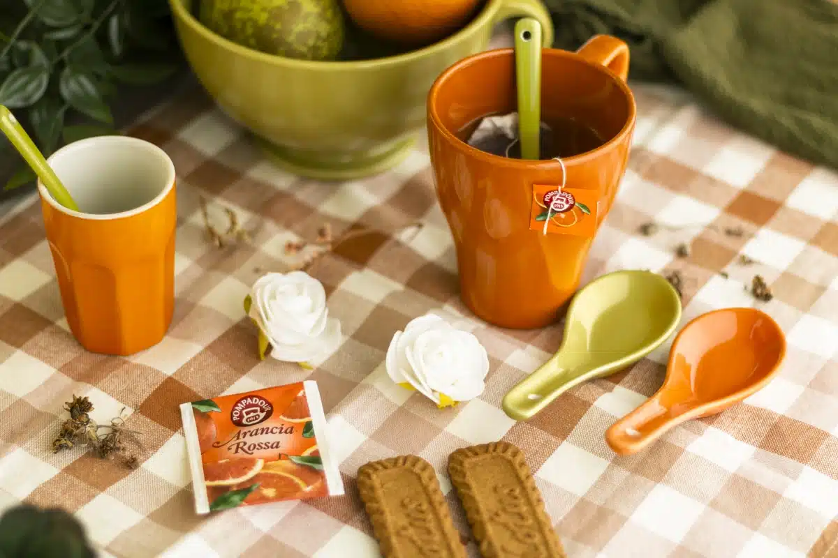 Een moodboard met een gezellige theetijd opstelling op een geruit tafelkleed. Te zien zijn een oranje mok met thee, een oranje beker, twee lepelhouders (een groene en een oranje), twee speculaas koekjes, een theezakje van Pompadour Arancia Rossa, en een kom met fruit op de achtergrond. Decoratieve witte rozen en droge takjes zijn verspreid over het tafelkleed.